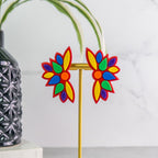 Colorful flower-shaped pride earrings on a stand with a plant and vase in the background.