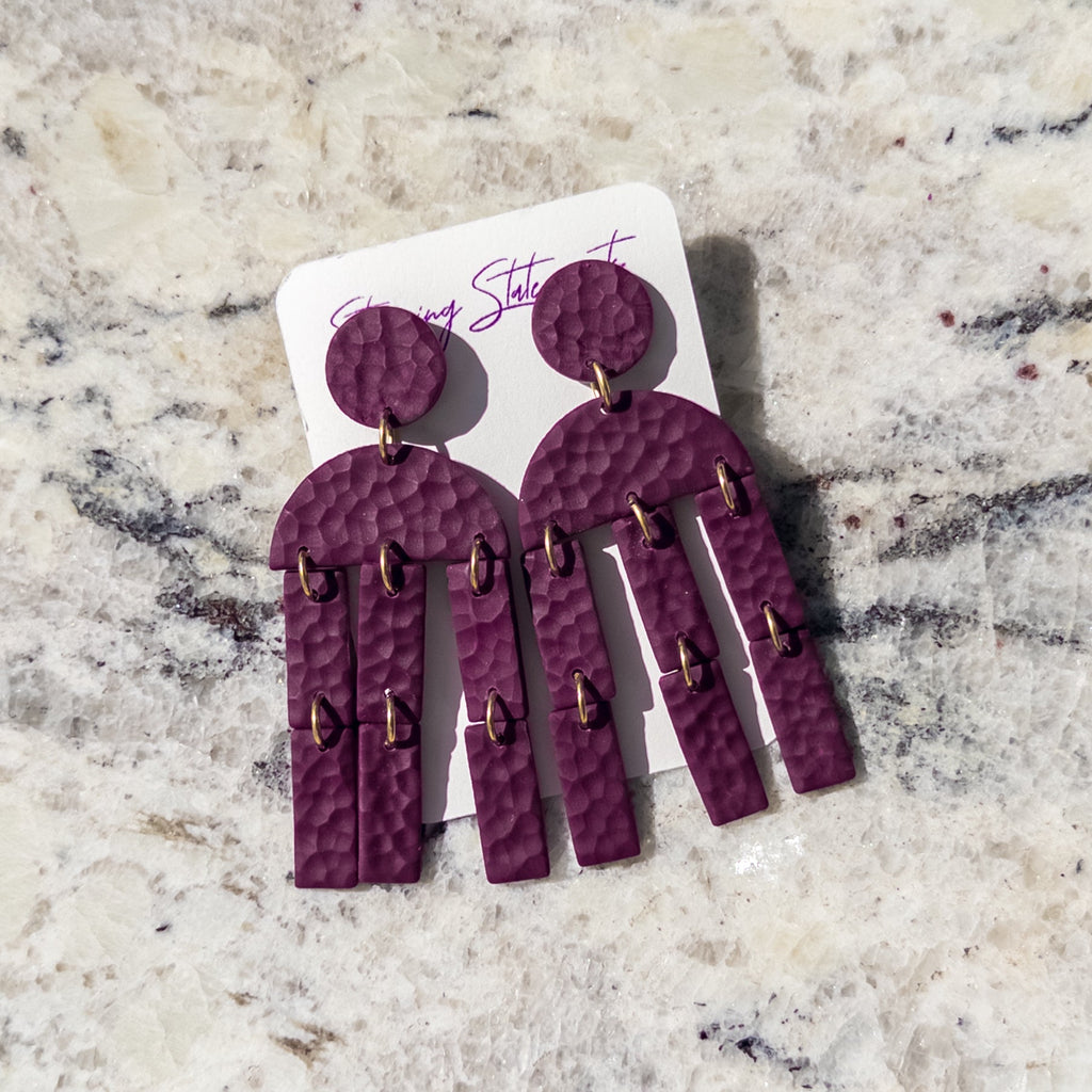 A pair of hammered purple statement earrings with gold-tone rings, displayed on a white backing card on a light beige marble surface.