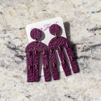 A pair of hammered purple statement earrings with gold-tone rings, displayed on a white backing card on a light beige marble surface.