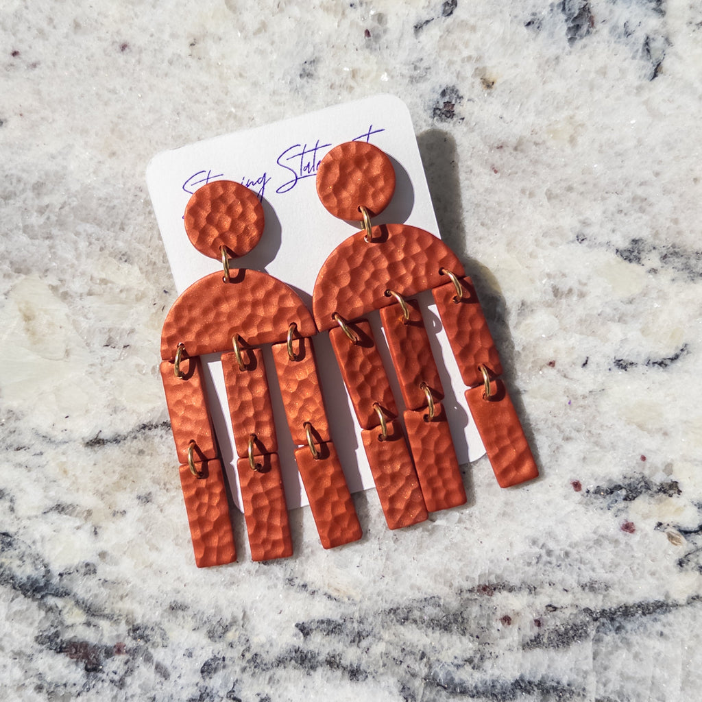 A pair of hammered terracotta orange drop earrings with small gold-tone ring accents, displayed on a white card over a light gray and white marble surface.