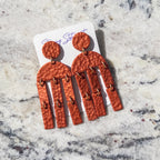 A pair of hammered terracotta orange drop earrings with small gold-tone ring accents, displayed on a white card over a light gray and white marble surface.
