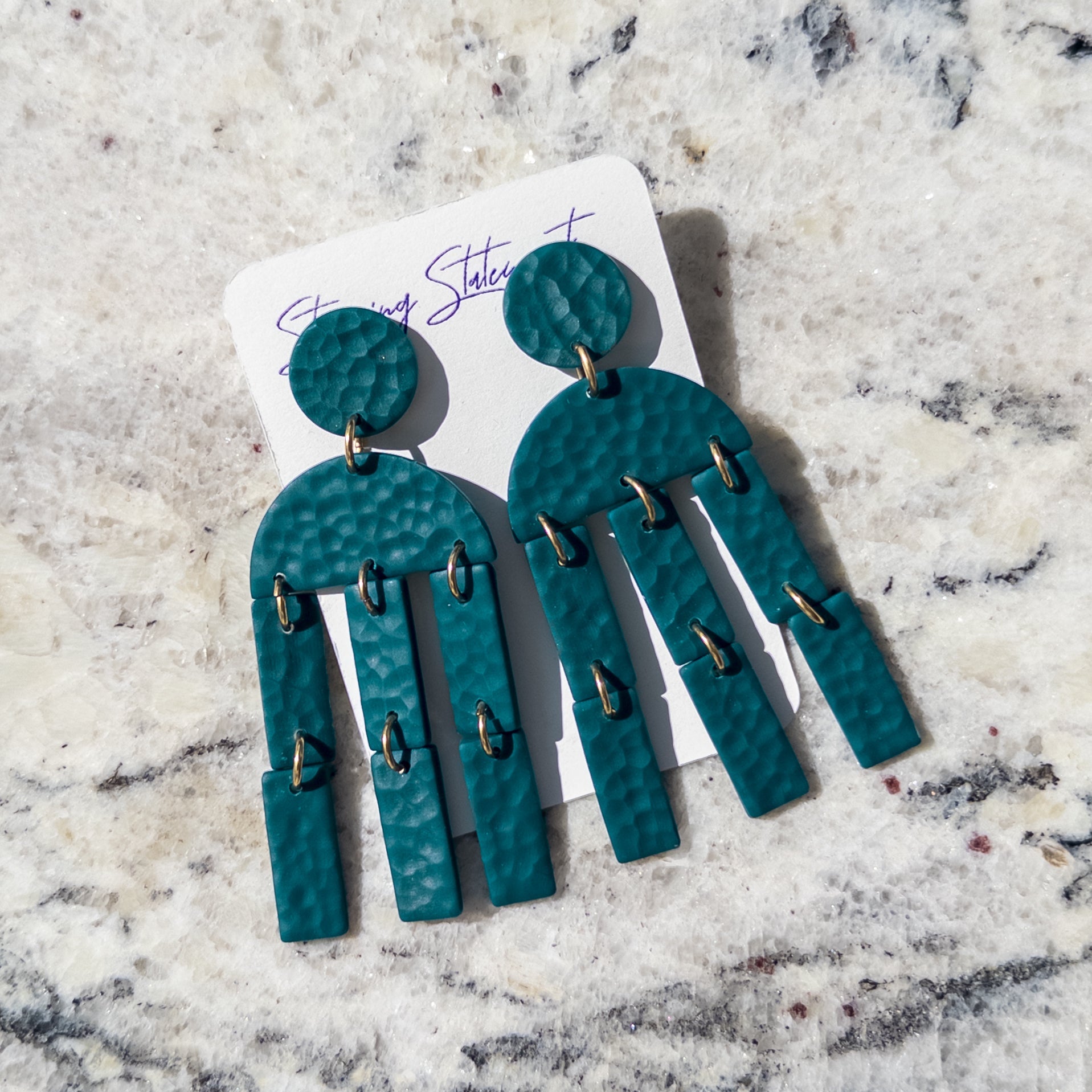 A pair of teal textured drop earrings with multiple dangling fringe sections and small gold-tone ring accents, displayed on a white card over a light marble surface.