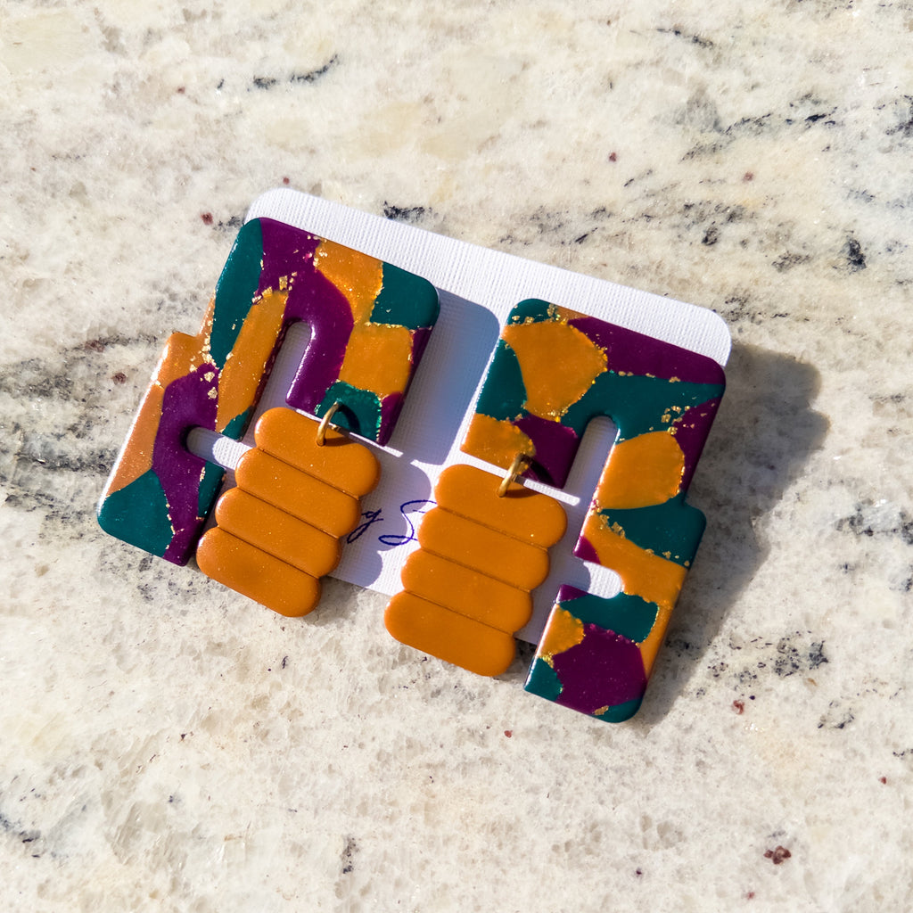 A pair of chunky geometric statement earrings with a marbled pattern of orange, purple, and teal, displayed on a white backing card over a light beige marble surface.