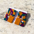 A pair of chunky geometric statement earrings with a marbled pattern of orange, purple, and teal, displayed on a white backing card over a light beige marble surface.