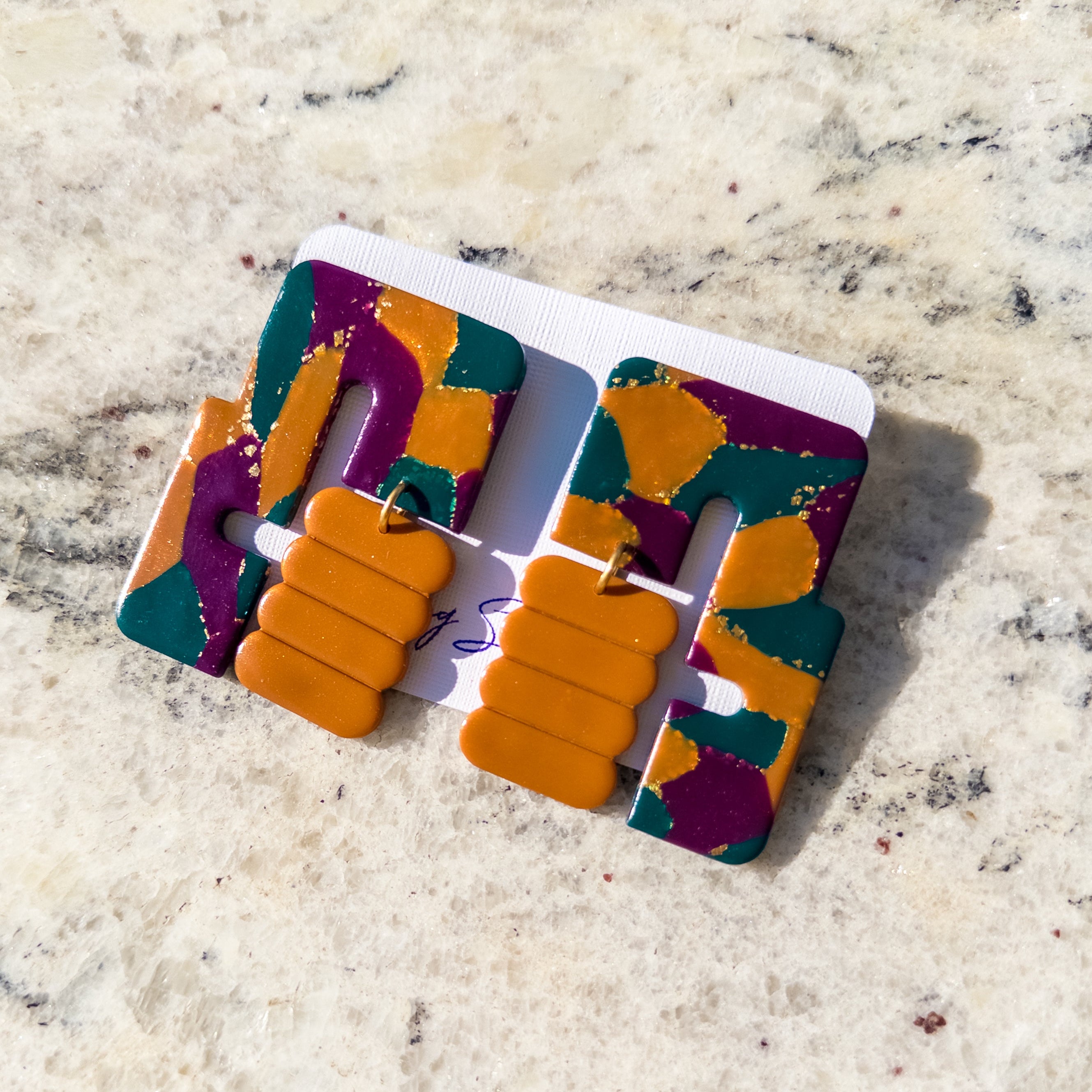 A pair of chunky geometric statement earrings with a marbled pattern of orange, purple, and teal, displayed on a white backing card over a light beige marble surface.