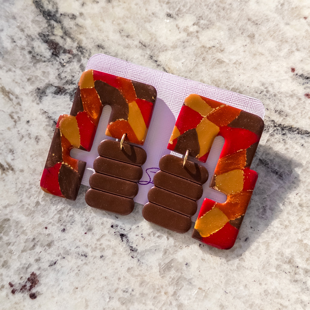 A pair of chunky geometric statement earrings displayed on a white card. The earrings feature a bold abstract pattern of red, orange, yellow, and brown shards on the outer arms, with three stacked brown rectangular plates connected by small gold rings in the center.