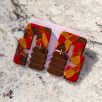 A pair of chunky geometric statement earrings displayed on a white card. The earrings feature a bold abstract pattern of red, orange, yellow, and brown shards on the outer arms, with three stacked brown rectangular plates connected by small gold rings in the center.