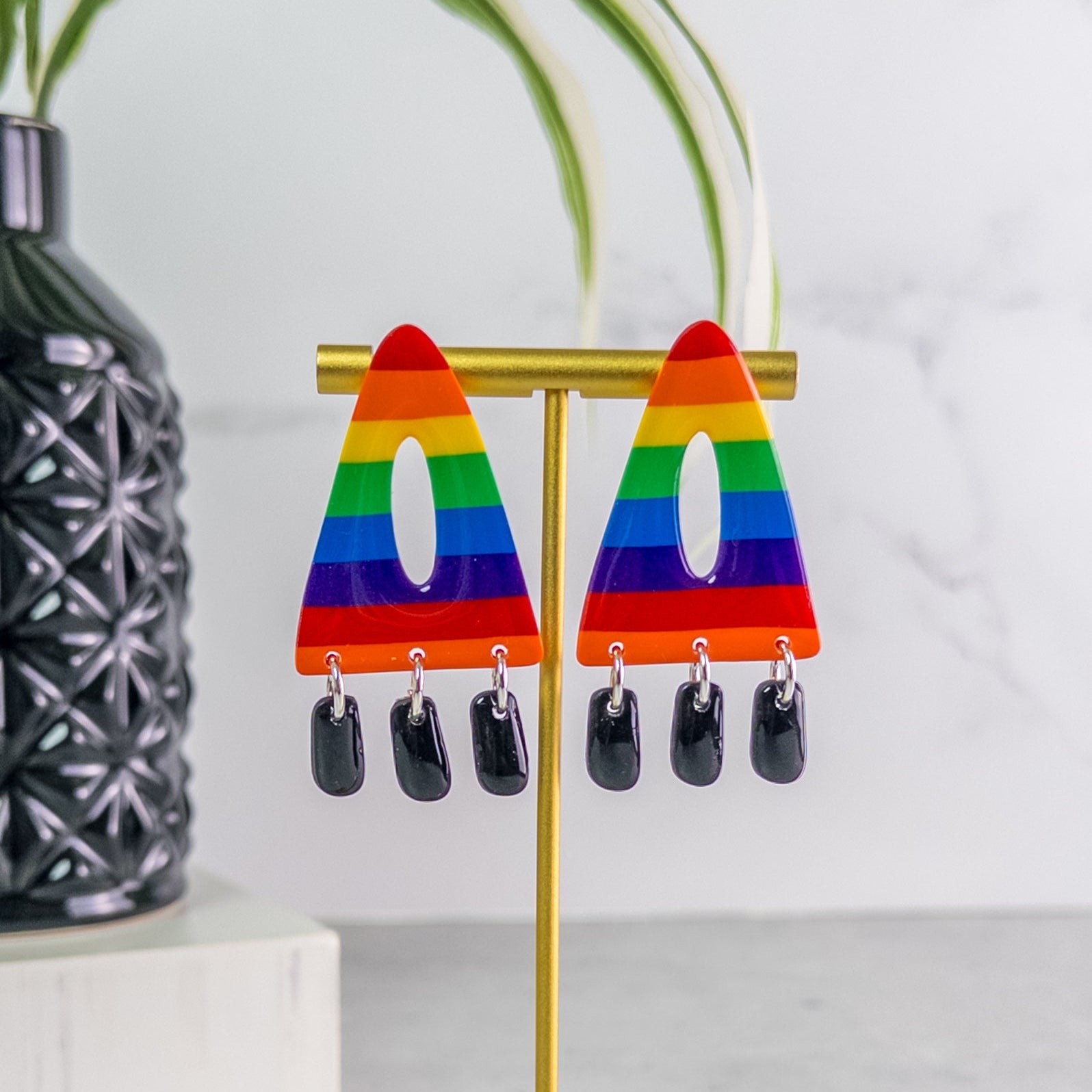 Rainbow pride  triangle earrings on a gold stand with a plant and vase in the background